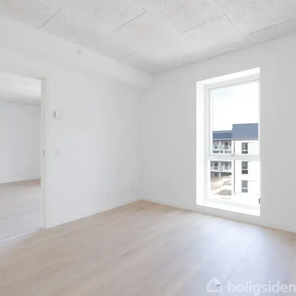 Et lyst værelse med trægulv står tomt. En åben dør fører til et tilstødende rum. Et stort vindue afslører moderne lejlighedsbyggeri udenfor.