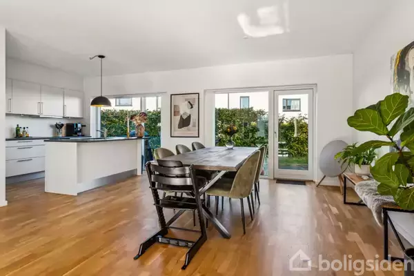 Spisebord med stole står i en lys stue med trægulv. En person står i baggrunden ved et moderne køkken med hvide skabe. Vinduer åbner til haven.