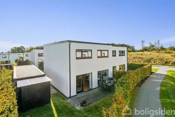 To-etagers hvidt hus med store vinduer står ved en brolagt terrasse med havemøbler. Omkranset af en hæk og placeret ved en snoet vej i et grønt villakvarter.