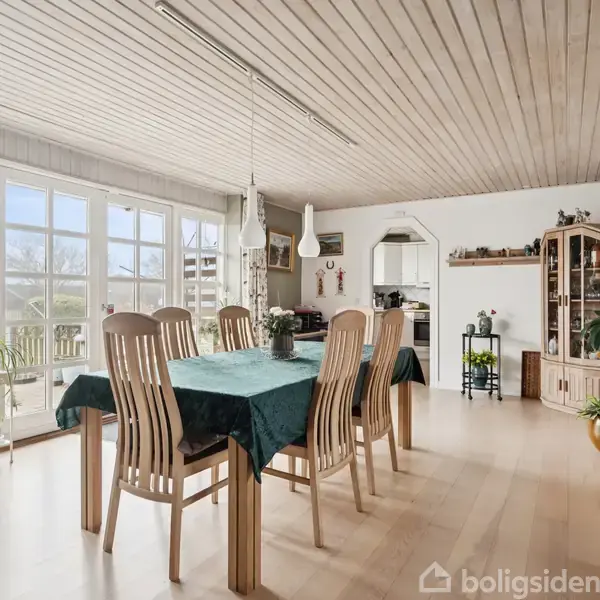 Spisebord står centralt dækket med grøn dug og seks stole omkring det. Stuen er lys med store vinduer, træloft og planter. Køkkenet ses i baggrunden.