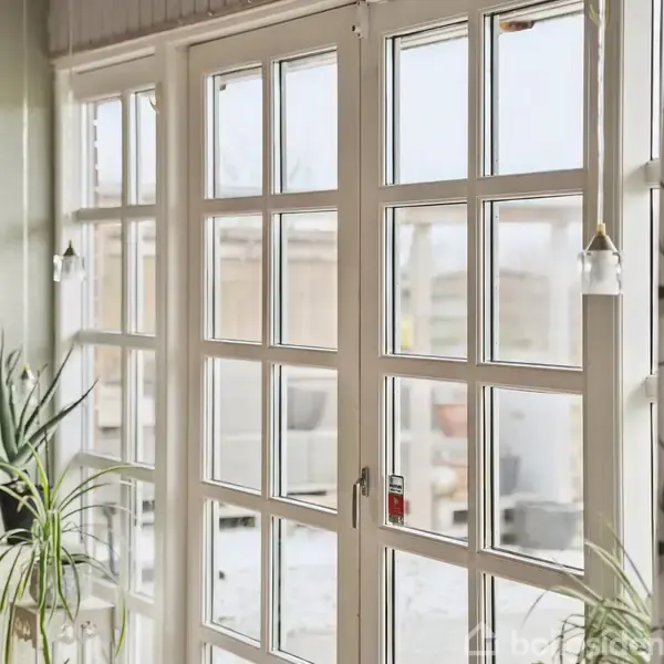 Et stort hvidt fransk vindue står åbent mod en udendørs scene. Hængende lamper og planter pryder rummet, omgivet af gardiner med blomstermønster. Vinterlys strømmer ind.
