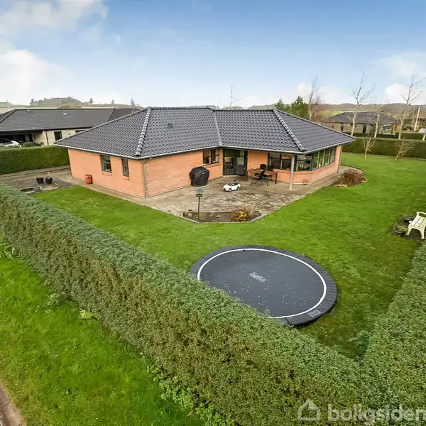 Et murstenshus står omgivet af en velplejet have med hække. En trampolin er placeret på græsplænen, og en terrasse er indrettet med møbler. Husene ses i et villakvarter.