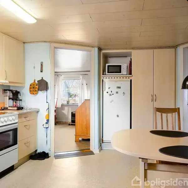 Køkken med komfur til venstre og spisebord til højre, omgivet af hvide skabe. En åbning fører til et tilstødende rum, og der hænger en sort lampe over bordet.