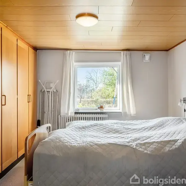 En dobbeltseng med hvidt sengetæppe står i et soveværelse med træloft. Til venstre er der store klædeskabe, og et vindue med udsigt til træer i baggrunden.
