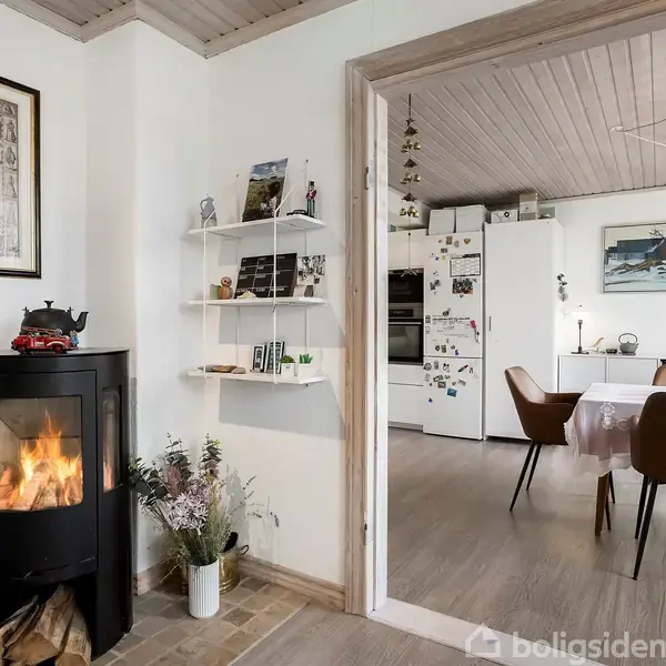 En brændeovn brænder i et hjørne af en stue med en kort hylde på væggen. I baggrunden ses et køkkenområde med spisebord og stole.