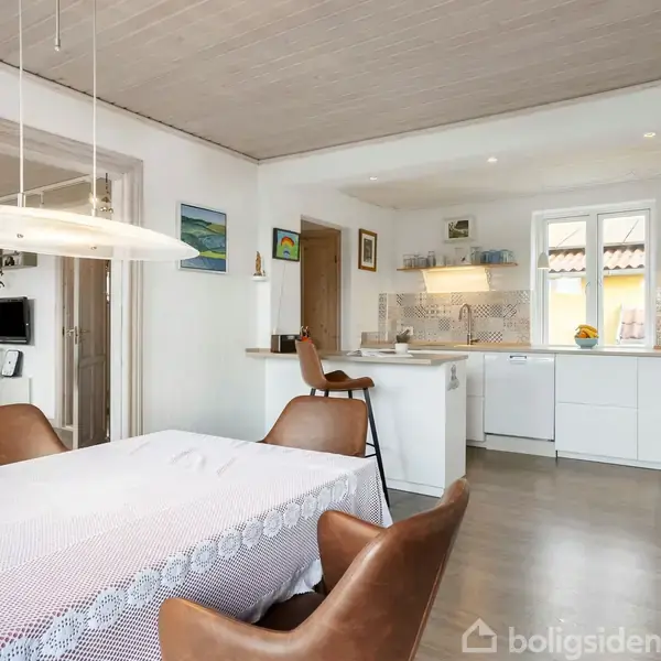 Spisebord dækket med dug står centralt, omgivet af brune stole i en åben planløsning, der fører til et lyst køkken med hvide skabe og åbne hylder, dekoreret med malerier på væggene.