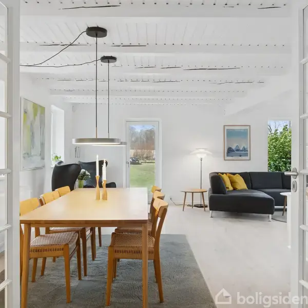 Et spisebord med træstole står midt i en lys stue. En sort sofa er placeret i baggrunden under et stort vindue, omkranset af hvide vægge og lofter med synlige bjælker.