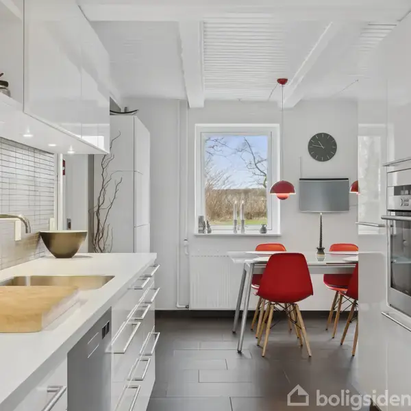 Et moderne køkken med hvide skabe har et spisebord med røde stole. En vægklokke hænger ved vinduet, der giver udsigt til et træ og græsplæne udenfor.