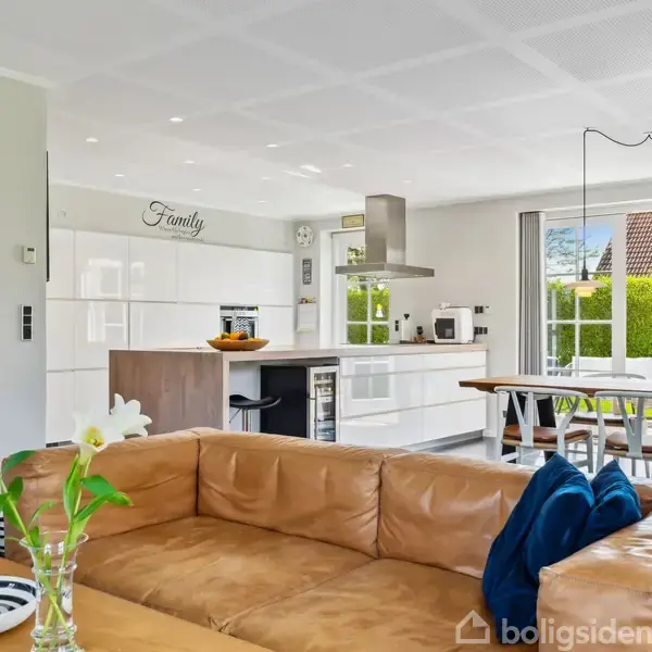 Brun lædersofa i stue vender mod køkken med hvide skabe. På væggen står "Family". Et spisebord med stole står ved siden af store vinduer, der giver udsigt til haven.