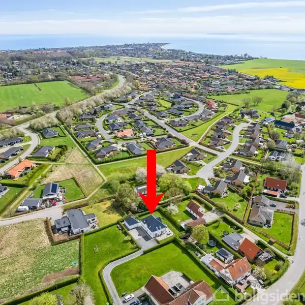 Et parcelhus er markeret med en rød pil. Det ligger i et villakvarter omgivet af grønne marker og nær kystlinjen i et åbent landskab.
