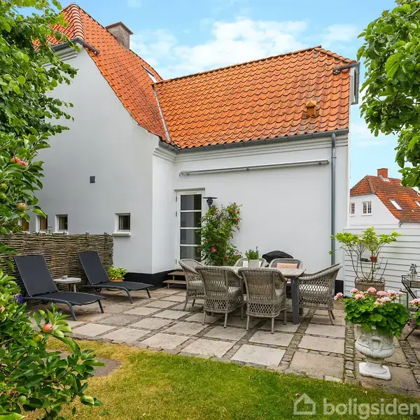 Havemøbler står på en flisebelagt terrasse ved et hvidt hus med rødt tegltag. To liggestole og et træbord med stole er omgivet af grønne buske og planter.