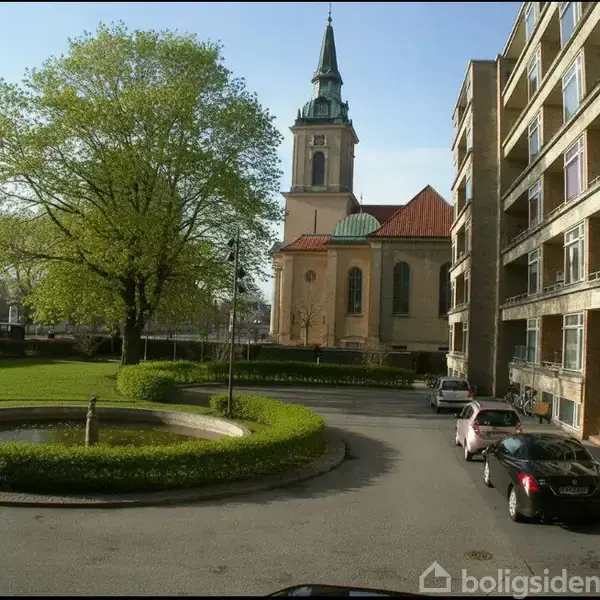 En høj, grøn træ står ved siden af en rund fontæne i en gårdhave. I baggrunden ses en kirke med et spir, omgivet af et boligkompleks til højre.