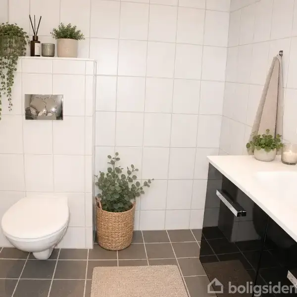 Et moderne badeværelse med hvide fliser. En væghængt toilet til venstre omgivet af planter i potter. Højre side har en sort vaskeskab med hvid vask og planter ovenpå.