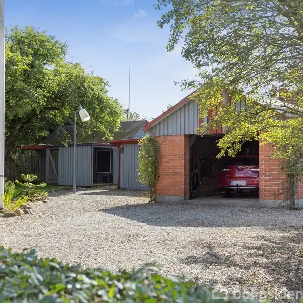 En rød bil står parkeret i en åben garage ved et rødt murstenshus. Omgivelserne er en grusindkørsel og grønne træer.