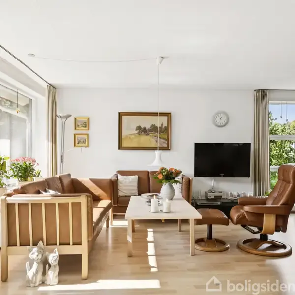 Stue med brun lædersofa og lænestol ved sofabord. Store vinduer giver lys. Vægdekorationer inkluderer maleri og ur. Potteplanter og bøger fylder rummet. Fladskærms-tv på væggen.