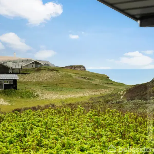 Et sort sommerhus står på grønne klitter med flere bygninger i baggrunden og havet i horisonten; udsigten ses gennem en ramme med en delvist overskyet blå himmel.