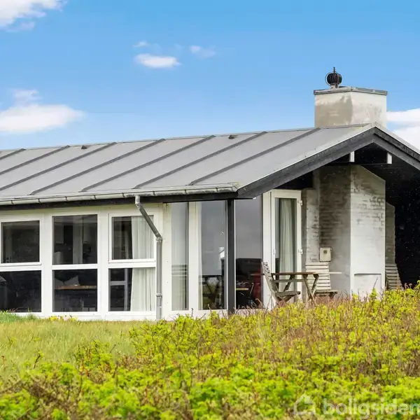 Et moderne sommerhus med glasfacade og sort tag står i en grøn, græsbevokset have. Ved huset står havemøbler foran en muret skorsten. Himmel med spredte skyer ses i baggrunden.