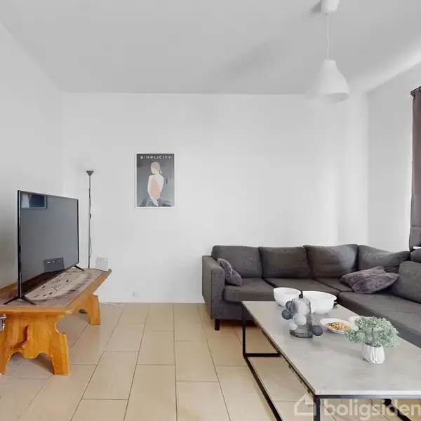 Stue med grå hjørnesofa, sofabord foran, og fjernsyn placeret til venstre på træbord. Hvid væg bagved med enkelt kunstværk. Loft til gulv gardiner i højre side.