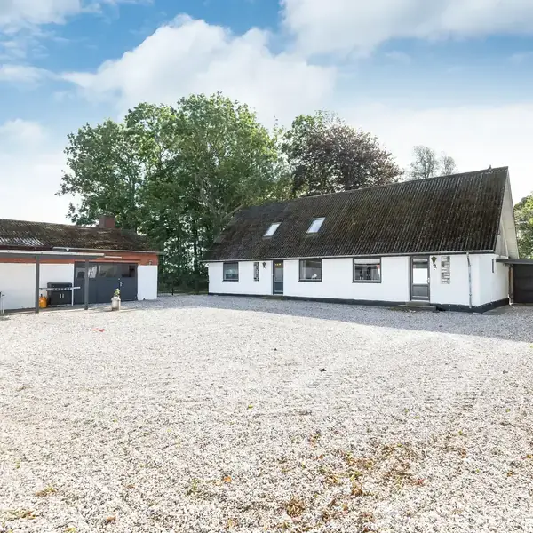 Hvid bygning med skrå tag, placeret på en gårdsplads med grusbelægning, omgivet af træer i baggrunden. Ved siden af ses endnu en bygning med terrasseområde.