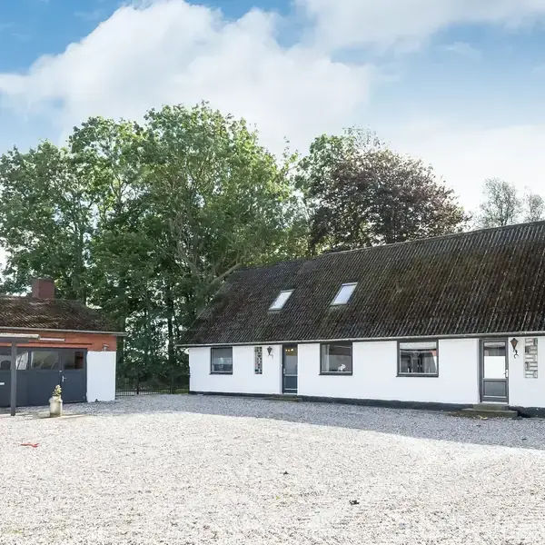Hus med hvid facade og skråtaget står stille på en åben plads omgivet af grus. Til venstre ses en carport. Træer i baggrunden beskriver naturmiljøet.