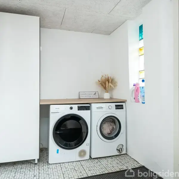 Vaskemaskine og tørretumbler står ved siden af hinanden under en hvid bordplade i et lyst vaskerum. Skabe og rengøringsmidler er i rummet, et vindue giver naturligt lys.