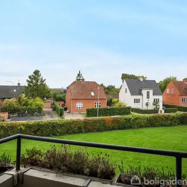 En balkon med blomsterkasser står stille med udsigt over en velholdt græsplæne, hvor der i baggrunden ses rækkehuse med rød og hvid facade.