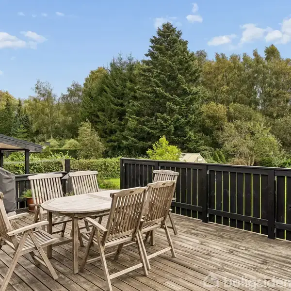 Udendørs træterrasse med et rundt spisebord og seks stole. Bordet er stationært, mens stolene er placeret omkring det. I baggrunden er der en grill og frodig skovudsigt.