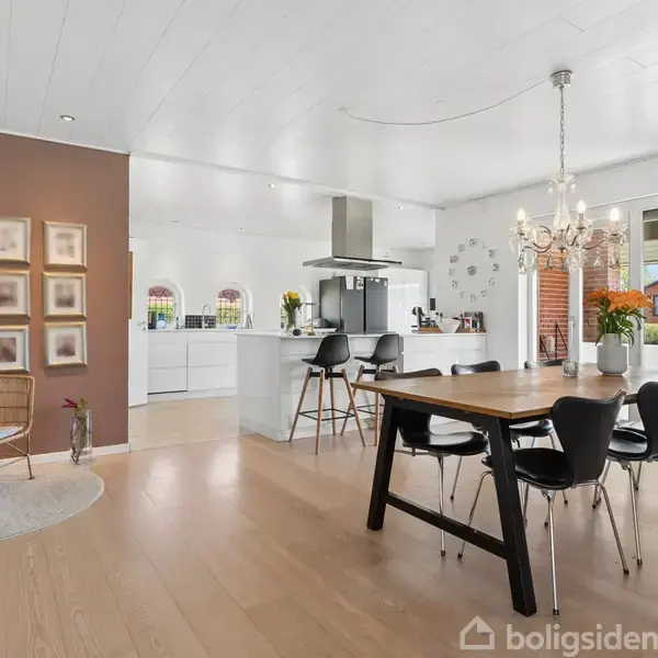 Et stort, lyst køkken-alrum med spisebord og stole i forgrunden. Køkkenø med barstole i baggrunden. En lysekrone hænger over bordet, og væggen er dekoreret med billeder.