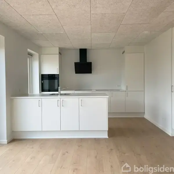 Et moderne køkken med hvide skabe, kogeplade og ovn integreret i væggen. Konteksten viser et lyst rum med trægulv og store vinduer, der giver naturligt lys.