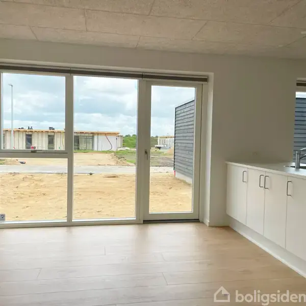 Flere store vinduer lader lys strømme ind i et moderne, lyst køkken med hvide køkkenskabe og en håndvask. Udenfor ses en sandet byggeplads med ufærdige bygninger i baggrunden.