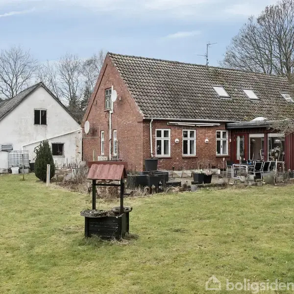 Et rødt murstenshus med skråt tag står i en have med græs. Ved siden af ses en hvid bygning, træer i baggrunden, og en brønd i forgrunden.