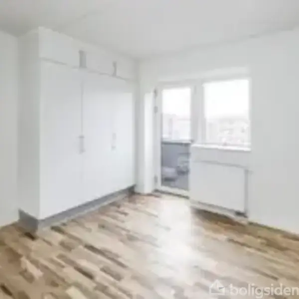 Værelse med garderobeskabe står stille i et lyst rum med trægulv. Dobbeltvindue og en radiator ses modsat entrédøren i baggrunden.
