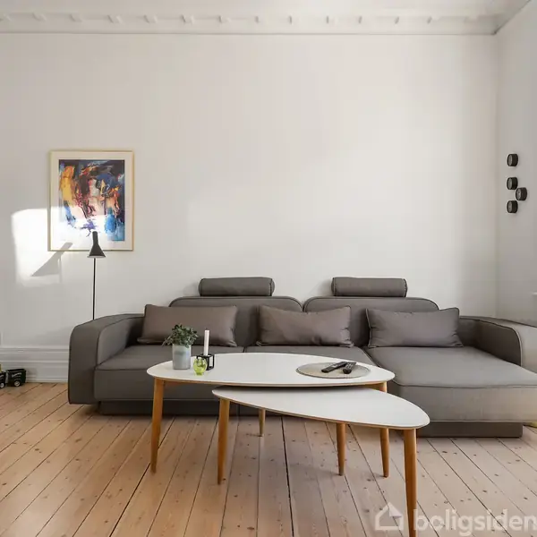Sofaen står i en lys stue på trægulv, omgivet af små borde med planter og en lampe. Et farverigt maleri hænger på væggen, og dagslys strømmer ind.