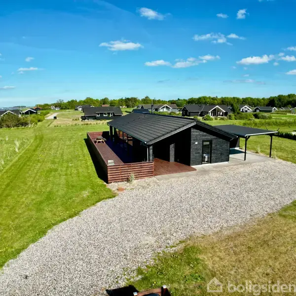 Et sort sommerhus står stille på en grusvej i en grøn eng nær en sø, under en klar blå himmel med spredte skyer.