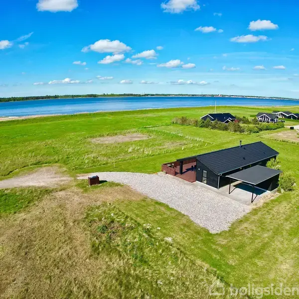 Et sort træhus med skråtag står på en stor, grøn græsplæne. Huset har en lang træterrasse med havemøbler og en grusbelagt indkørsel i et åbent sommerhusområde.