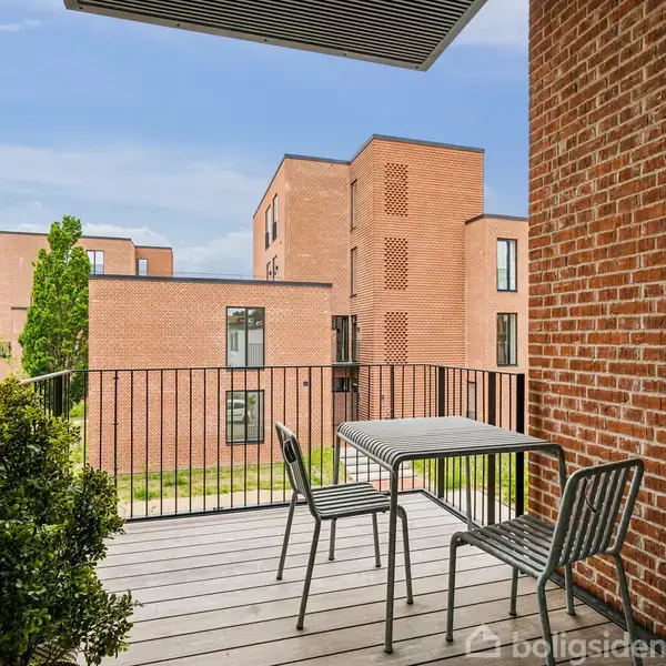Terrasse med to stole og et bord står stille mod en baggrund af moderne, røde murstensbygninger med flere etager. Grønne planter tilføjer et naturligt præg.