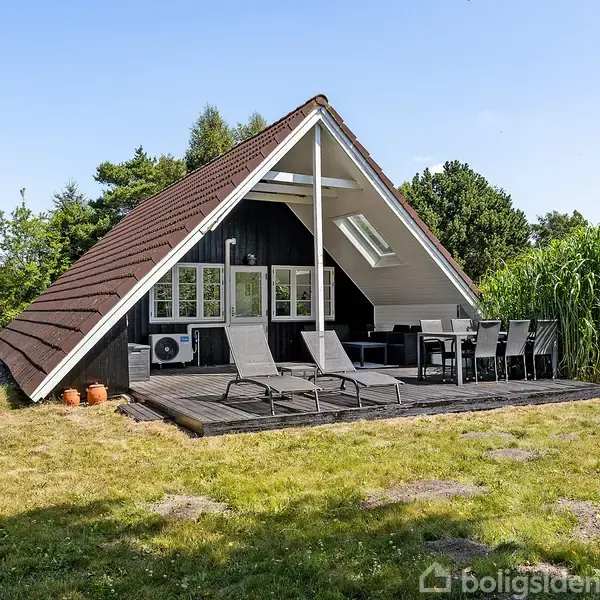 A-formet sommerhus med en træterrasse, hvor der står liggestole og et spisebordssæt. Omgivet af en grøn have og træer, under en klar blå himmel.