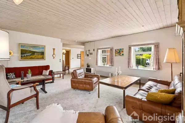 Stue med brune lædersofaer og et sofabord, omgivet af hvide vægge og træloft. Møbler står på et lyst tæppe. Vinduer viser en grøn have udenfor. Malerier hænger på væggene.