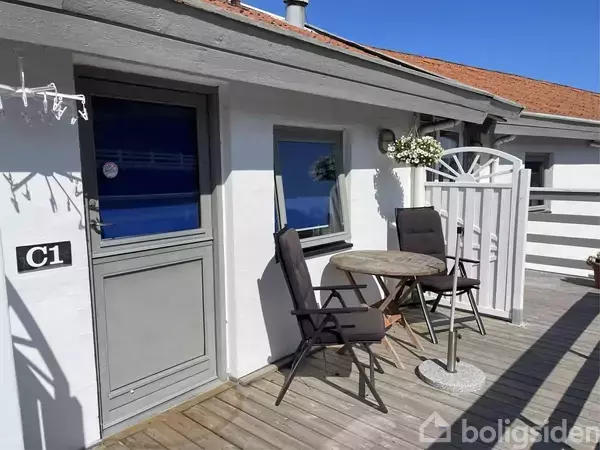 To stole og et rundt bord står på en terrasse foran en dør mærket "C1" til et hvidt hus med et træhegn. En hængende blomsterkurv pynter væggen.