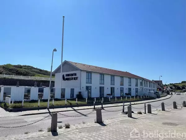 En lang bygning med skiltet "FERIECENTER HARBOØRE KUR" står i sollys ved en vej og fortov. To biler ses parkeret, og bakker danner baggrunden. Skinnende blå himmel over stedet.