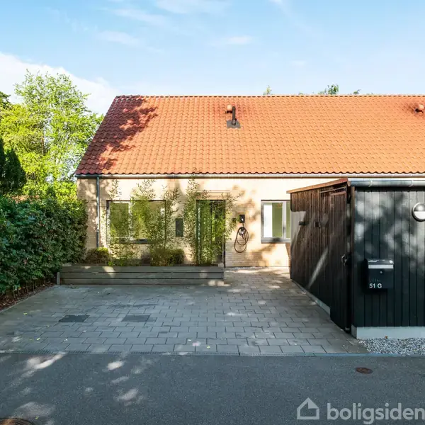 Et halvandetplans rækkehus med rød tegltag står stille under blå himmel, omgivet af grønne buske. En brolagt indkørsel fører til et sort skur og en postkasse mærket "51G".