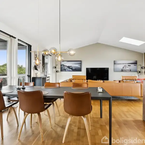 Et spisebord med seks stole står i et moderne, lyst rum; en sofa og fladskærm ses i baggrunden. Store vinduer giver udsigt til grønne omgivelser. Lampen hænger over bordet.