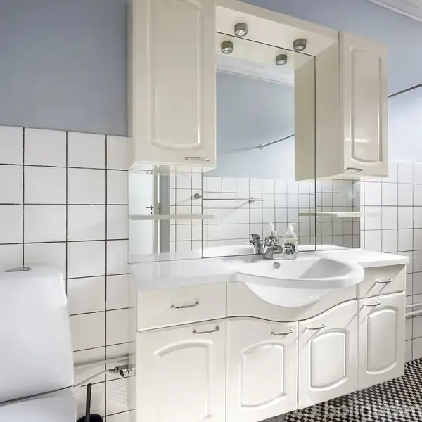 Badeværelsesvask med hvidt skab og spejl, placeres mod en væg med hvide fliser. Til venstre står en toiletstol, mens der til højre kan ses et tørrestativ og radiator.