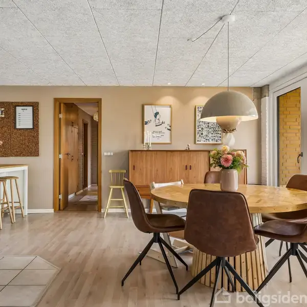 En rund spisebord med brune stole står i en lys stue. En moderne brændeovn opvarmer rummet. Store vinduer til venstre lader masser af lys ind. Vægdekorationer ses i baggrunden.