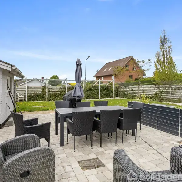 Terrassemøbler står opstillet omkring et bord på en flisebelagt terrasse. En parasol er placeret i midten. Et boligområde med hække og et hus ses i baggrunden.