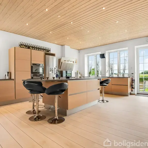 Et moderne køkken med træloft, køkkenø med tre sorte barstole og indbyggede ovne. Store vinduer og glasdøre giver udsigt til en grøn have. En træplankegulv skaber et lyst rum.