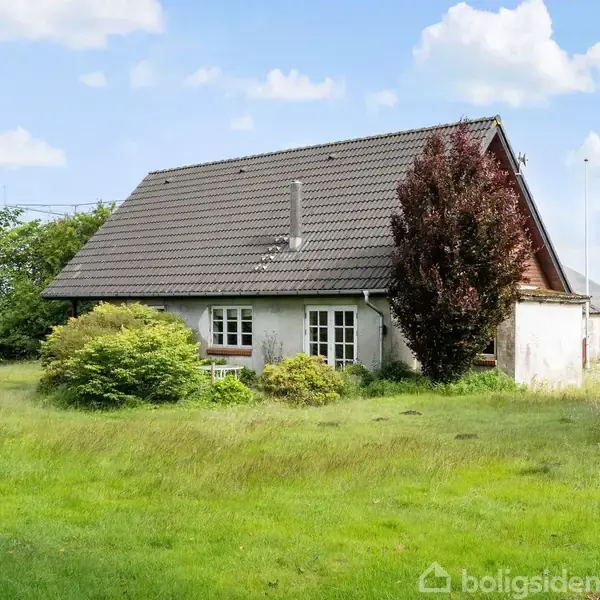 Et hvidt hus med mørkt tag står blandt grønne buske og en velplejet græsplæne. Skyfri himmel og træer danner en landlig baggrund.