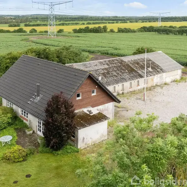 En gård med et mørkt tag står ved en grusplads omgivet af grønne marker og buske. En lille, hvid bord står i haven tæt på huset.