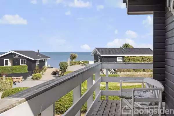 En balkon med en bænk vender ud mod en udsigt over havet. I forgrunden ses sorte huse og grøn vegetation med himlen i baggrunden.