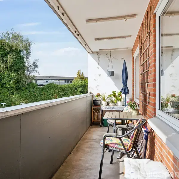 Balkon med stole og bord, overskygget af en parasol. Omgivet af murstensvægge og planter, med udsigt til en have og flere træer.